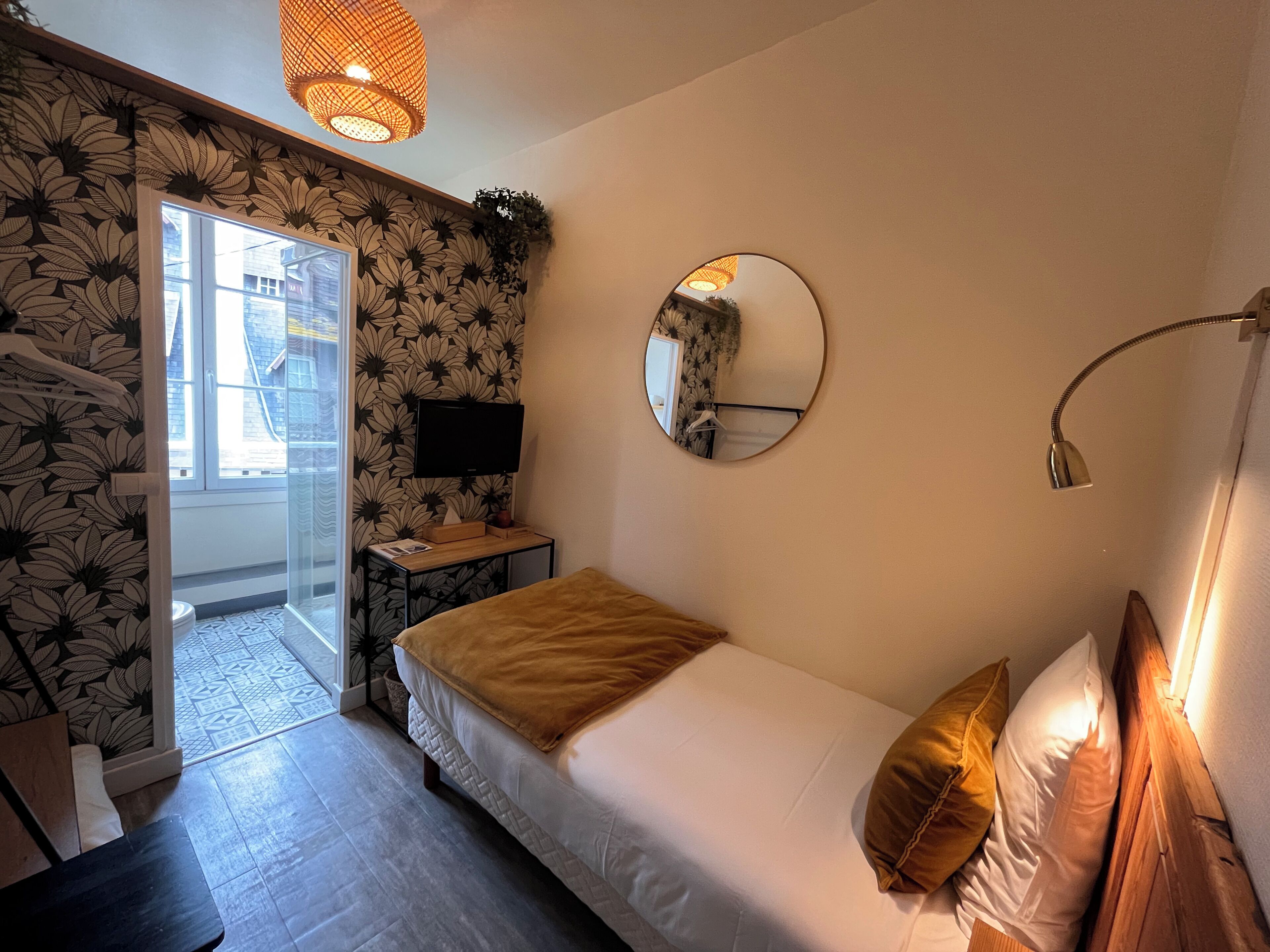A bedroom with a bed, a round mirror, a bedside lamp, a TV, and a window with a view of buildings.