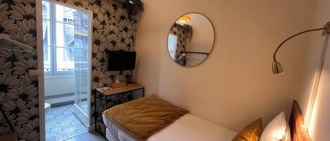 A bedroom with a bed, a round mirror, a bedside lamp, a TV, and a window with a view of buildings.