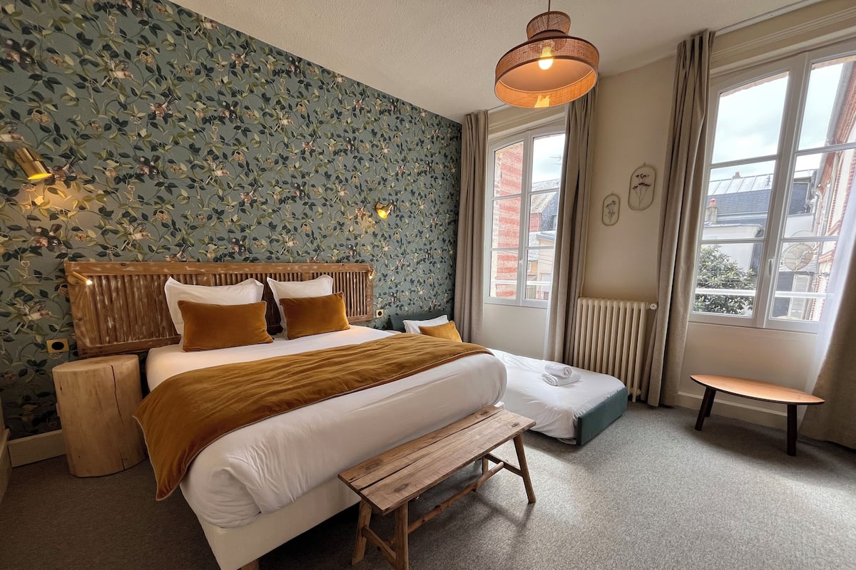 Een slaapkamer met een groot bed, een houten bankje, een klein tafeltje en een raam met gordijnen.