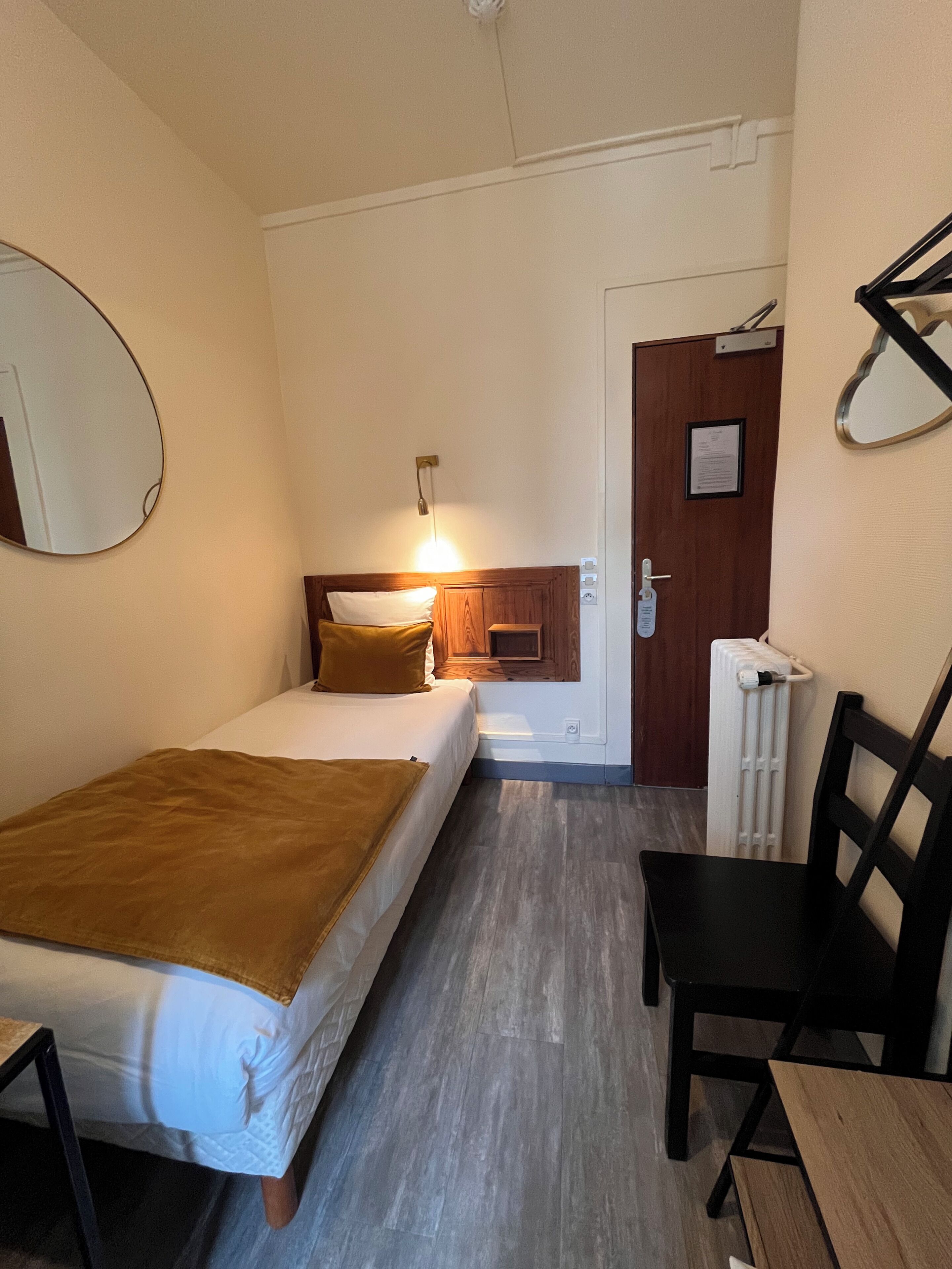 A hotel room with two beds, a wooden headboard, a radiator, a chair, and a door with a sign.