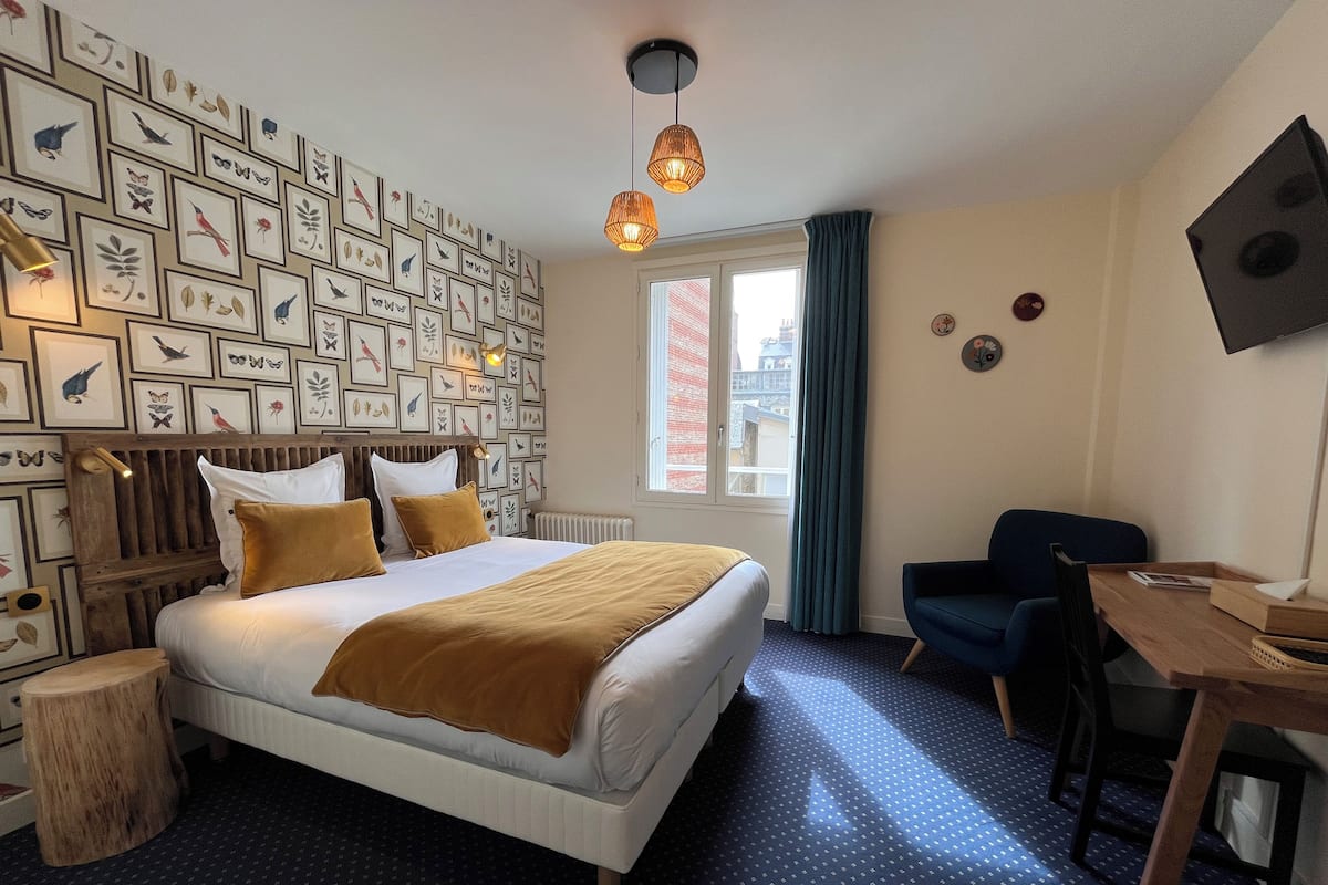 Een hotelkamer met een bed, een bureau, een stoel, een raam met gordijnen en een muur versierd met ingelijste foto's.
