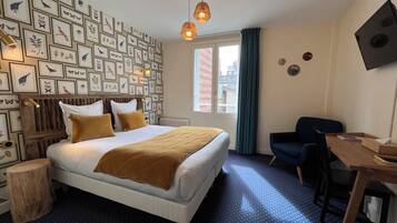 A hotel room with a bed, a desk, a chair, a window with curtains, and a wall decorated with framed pictures.