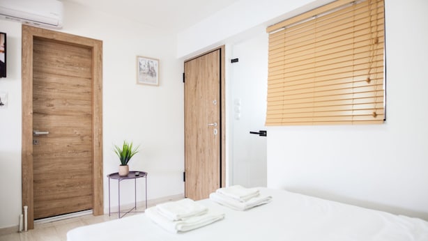 A bedroom with a bed, a wooden door, a small round table with a plant, and a window with blinds. - Spacious 2 level Brand New apt GTD3194 (Athens)