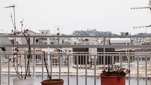 A balcony with potted plants, overlooking a cityscape with industrial buildings and a distant view of a historical site. - Spacious 2 level Brand New apt GTD3194 (Athens)