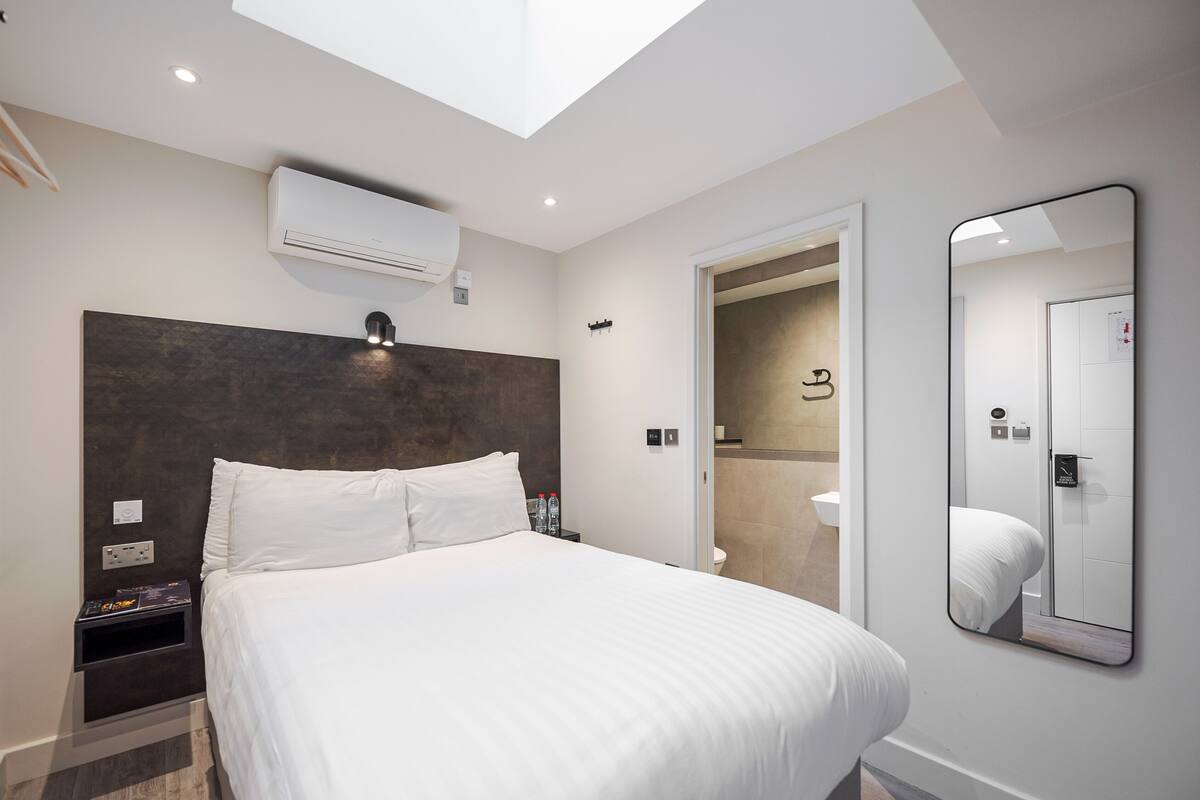 A modern hotel room with a large bed, a skylight, a wall-mounted air conditioner, a bedside table with a lamp, and a large mirror.
