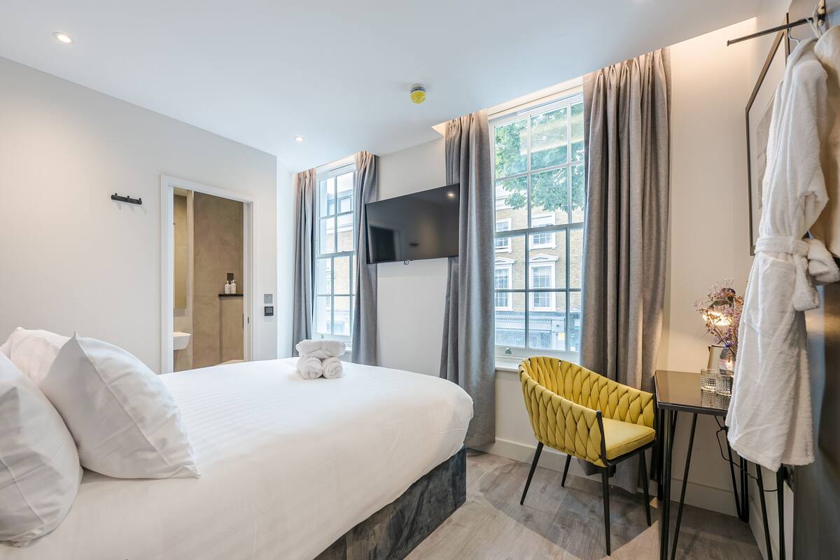 A modern hotel room with a large bed, a flat-screen TV, a small table with a chair, and a window with curtains.