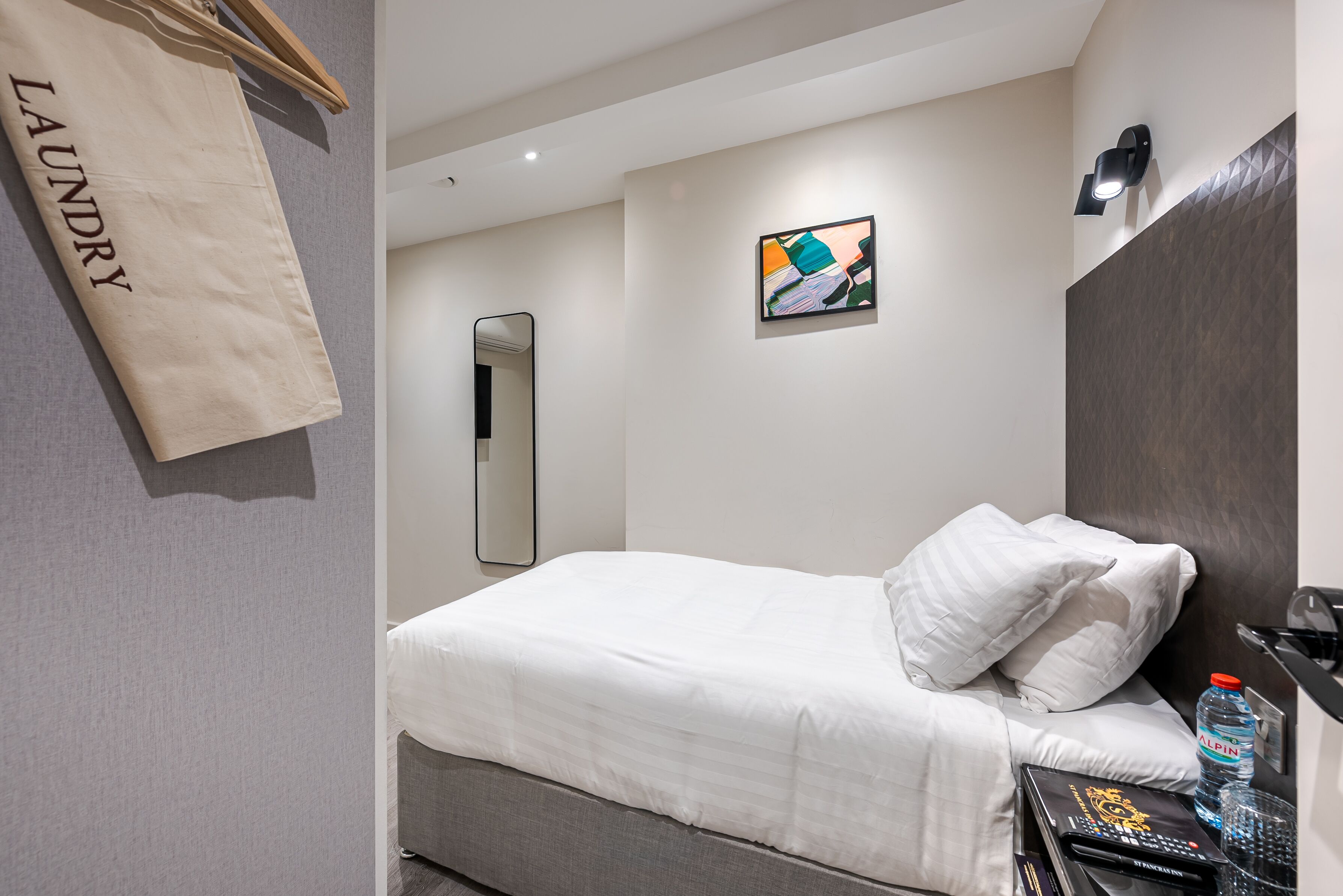 Een netjes opgemaakt bed met witte lakens, een klein nachtkastje met een waterfles, een afstandsbediening en een decoratief object. Een handdoekhouder met een 'laundry'-bord hangt aan de muur, en er is een klein abstract schilderij zichtbaar boven het bed.
