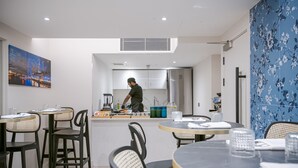 Un comedor moderno con mesas redondas y sillas negras, una cocina al fondo y un cuadro de un paisaje urbano en la pared.