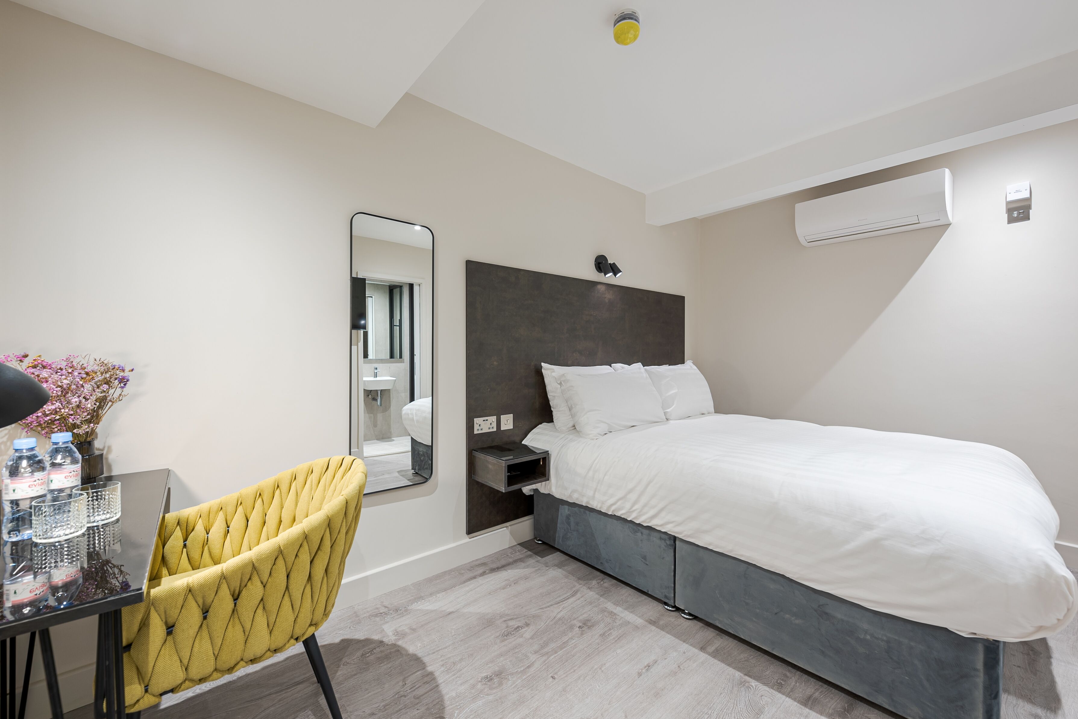 A modern hotel room with a bed, bedside table, chair, and a mirror.
