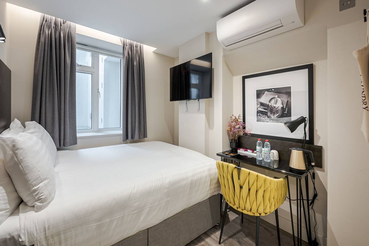 A hotel room with a bed, a desk with a chair, a flat-screen TV, an air conditioner, and a framed picture on the wall.