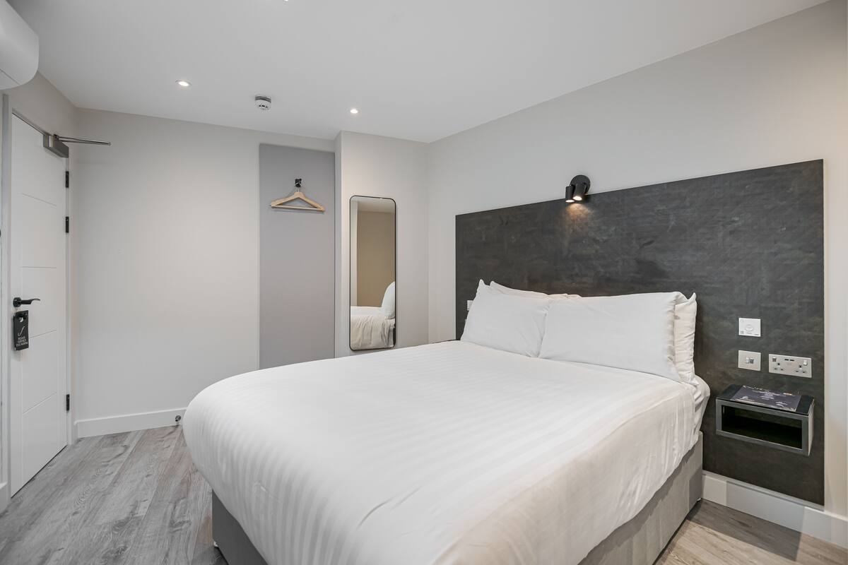 A modern hotel room with a large bed, a bedside table, a wall-mounted light, and a door with a handle.