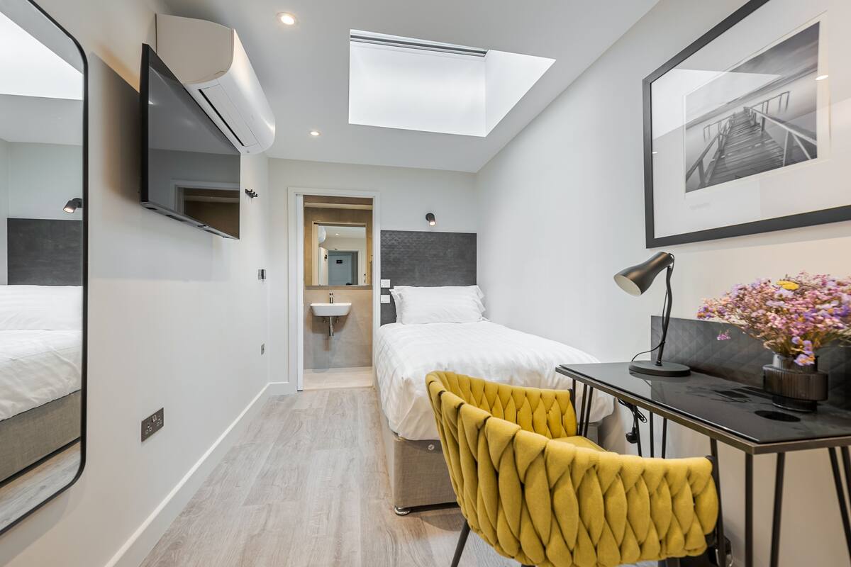 A modern hotel room with a bed, a desk with a chair, a mirror, a TV, and a skylight.