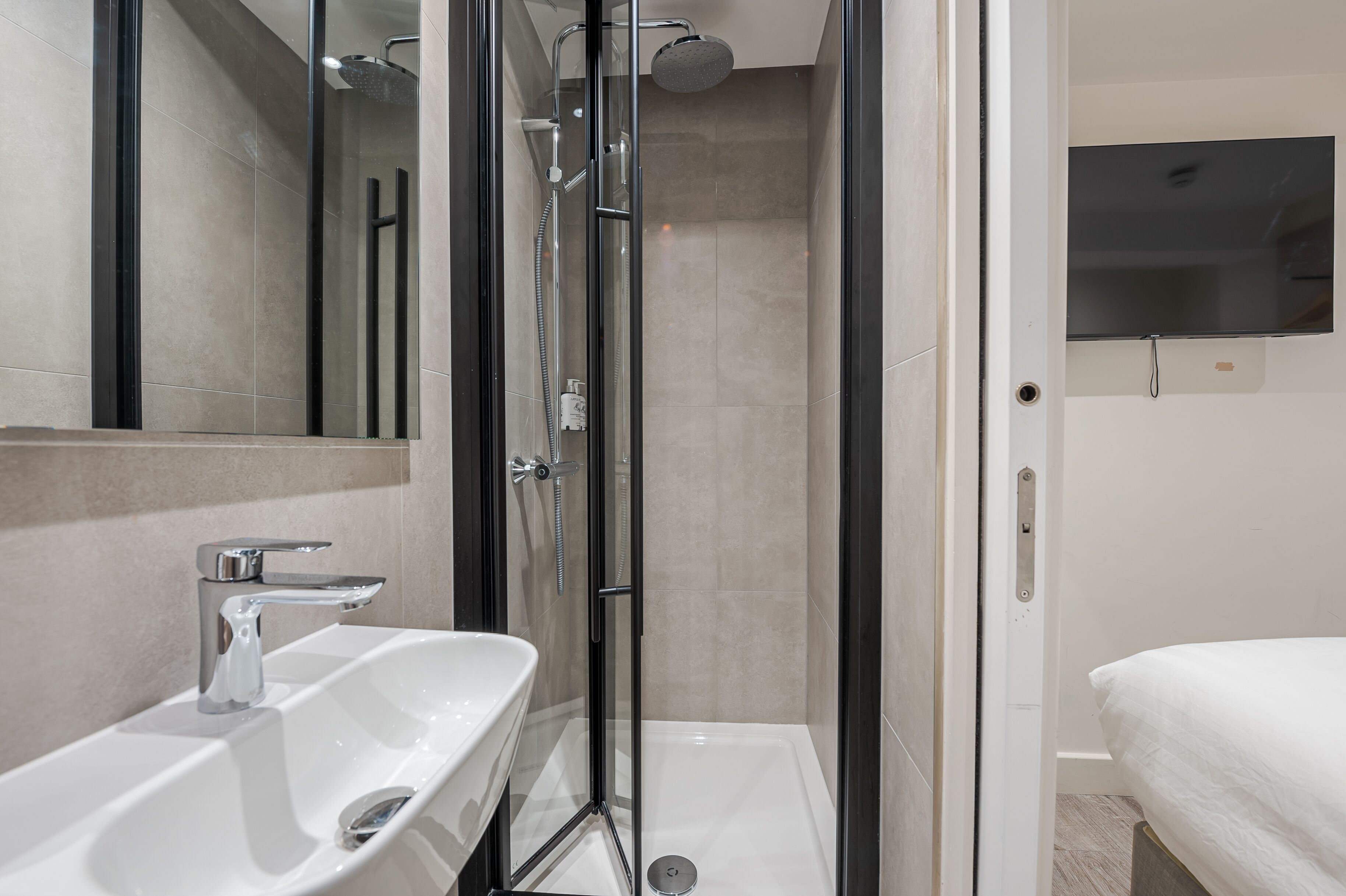 A modern bathroom with a glass-enclosed shower, a white sink, and a flat-screen TV mounted on the wall.