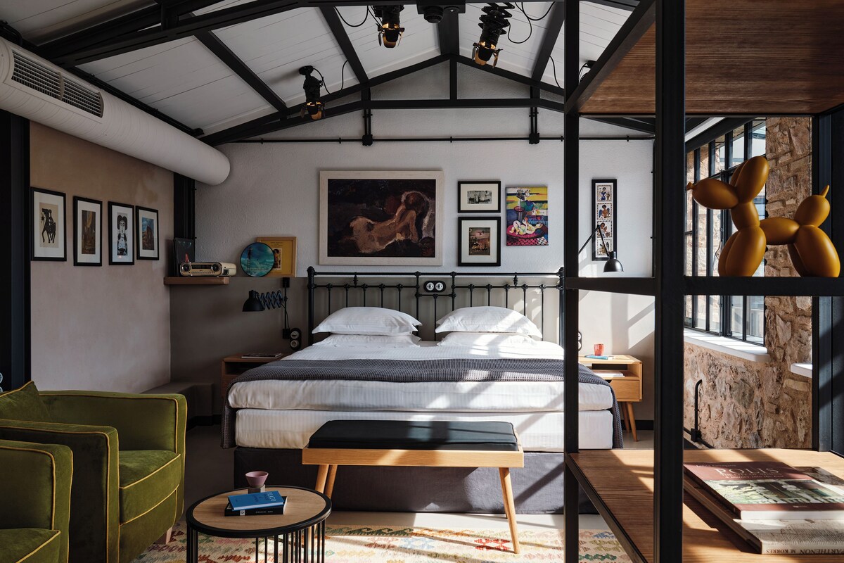 A modern bedroom with a large bed, a green armchair, a small round table, and a colorful rug.
