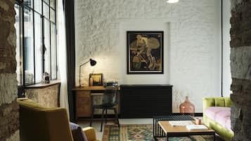A cozy room with a stone wall, a window, a desk with a lamp, a chair, a sofa, and a patterned rug.