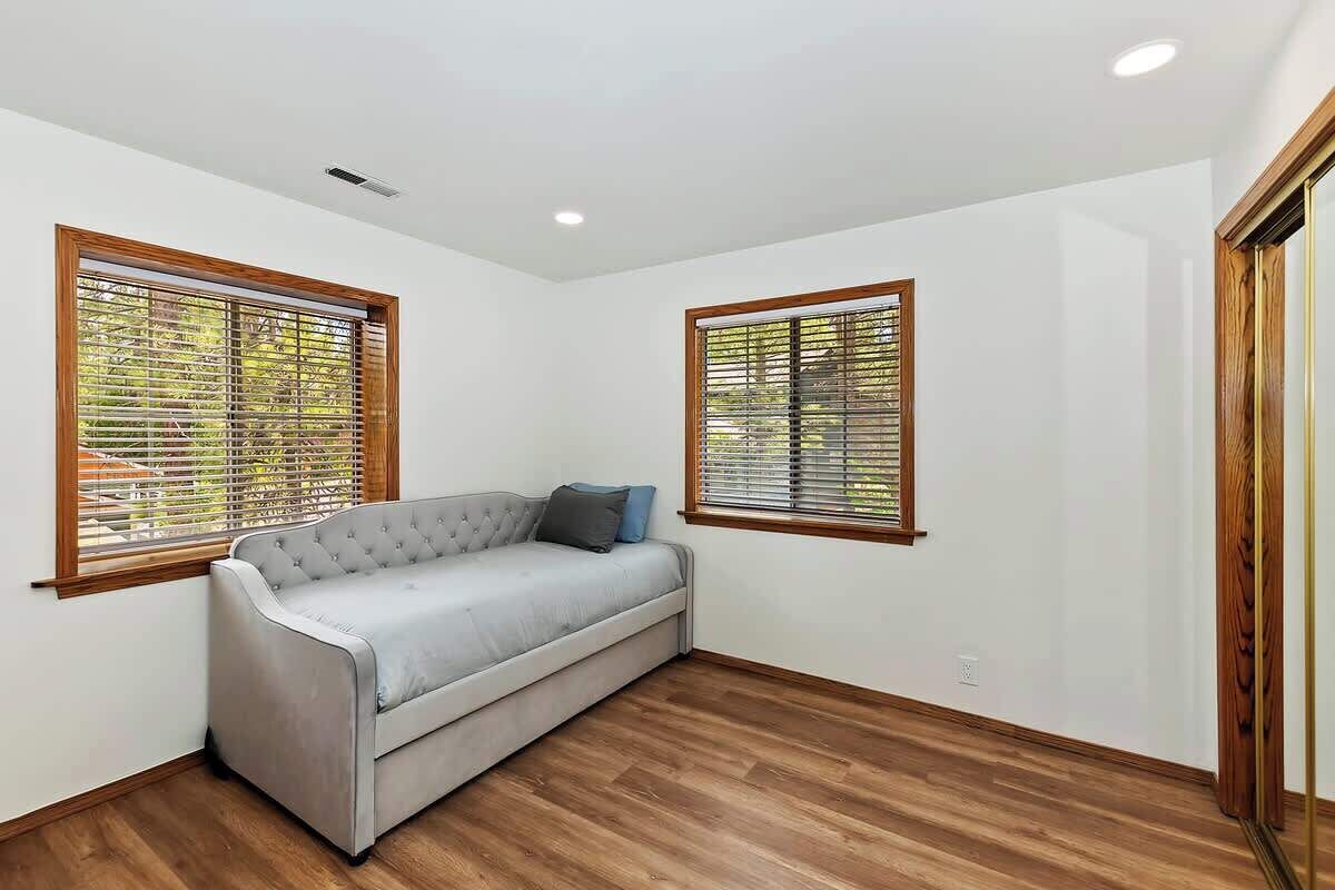 A room with a grey sofa, wooden flooring, and two windows with blinds.