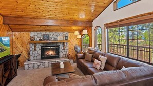 Un intérieur de chalet chaleureux avec une cheminée en pierre, un canapé en cuir et de grandes fenêtres offrant une vue sur l’extérieur.