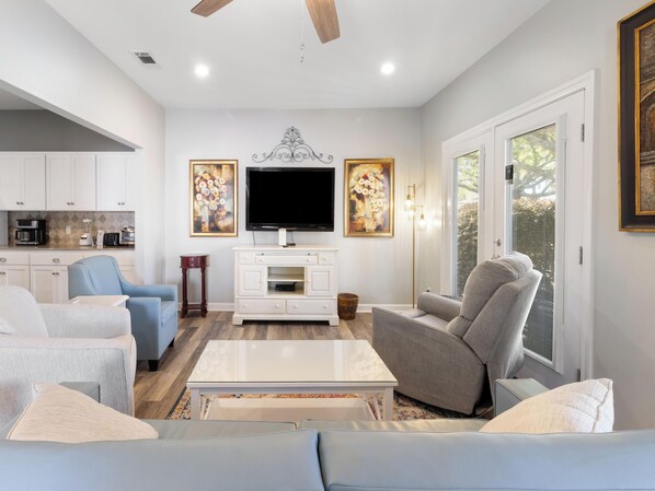 A modern living room with a flat-screen TV, two armchairs, a sofa, a coffee table, and a kitchen area in the background.