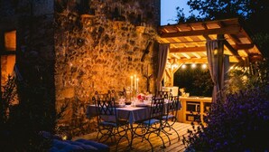 An outdoor dining area with a stone wall backdrop, wooden deck, and a pergola with string lights. - Romantic stay with spa, sauna and home cinema (Beaumontois-en-Périgord)