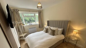 A bedroom with a large bed, a bedside table with a lamp, a radiator, a window with curtains, and a TV mounted on the wall. - Charming 2-bed Apartment in Danbury, Essex (Chelmsford)