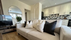 A living room with a beige sofa, a flat-screen TV, and a kitchen in the background. - Charming 2-bed Apartment in Danbury, Essex (Chelmsford)