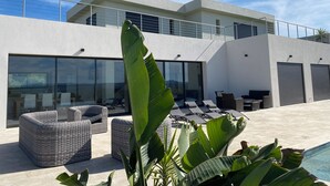 A modern outdoor seating area with wicker furniture, large windows, and a pool. - Beautiful new housse with splendid view and big pool. (Cotignac)