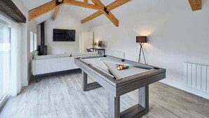 Una mesa de billar moderna en una habitación amplia con techo alto y vigas a la vista, chimenea y un ventanal que proporciona luz natural.