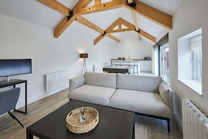 Un salon moderne avec un canapé, une table basse et un téléviseur. Au fond, on aperçoit une cuisine avec une table à manger et des chaises.