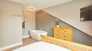 A modern bedroom with a large bed, a wooden dresser, a built-in bench, a wall-mounted TV, and a freestanding bathtub. - Host & Stay | The Old Jail Barber's Shop (Scarborough)