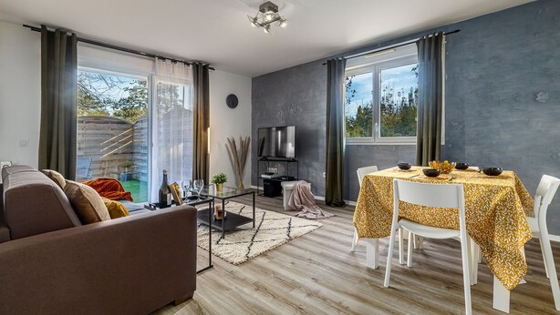 Ein modernes Wohnzimmer mit einer Couch, einem Esstisch mit Stühlen, einem Fernseher und großen Fenstern mit Vorhängen.