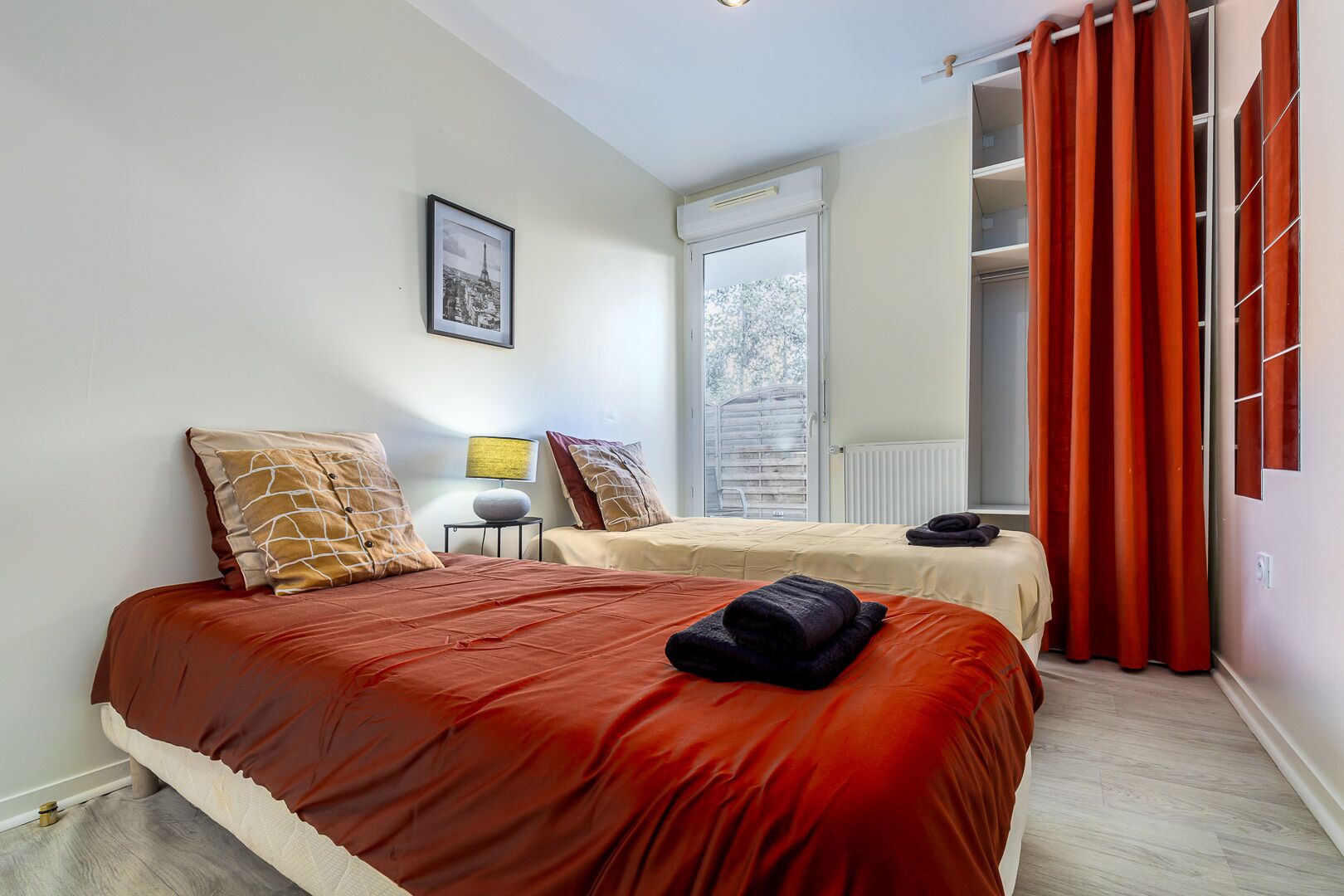 A hotel room with two beds, a window with a view, a framed picture on the wall, and a red curtain.
