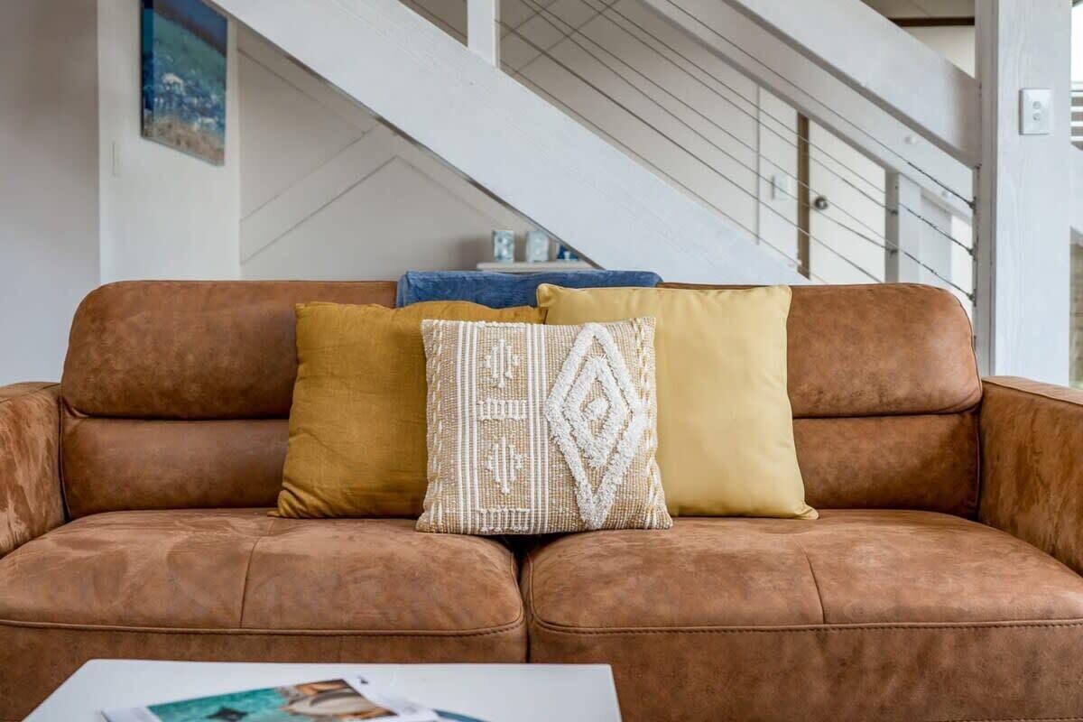 A brown leather sofa with three cushions in a modern living room with a staircase in the background.