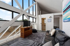 Avoin, loft-tyylinen huone, jossa on suuri ikkuna, puinen TV-taso, musta nahkasohva ja tikkaat.