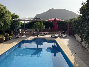 Ein Swimmingpool mit Liegestühlen und Sonnenschirmen, umgeben von Topfpflanzen und Blick auf einen Berg.