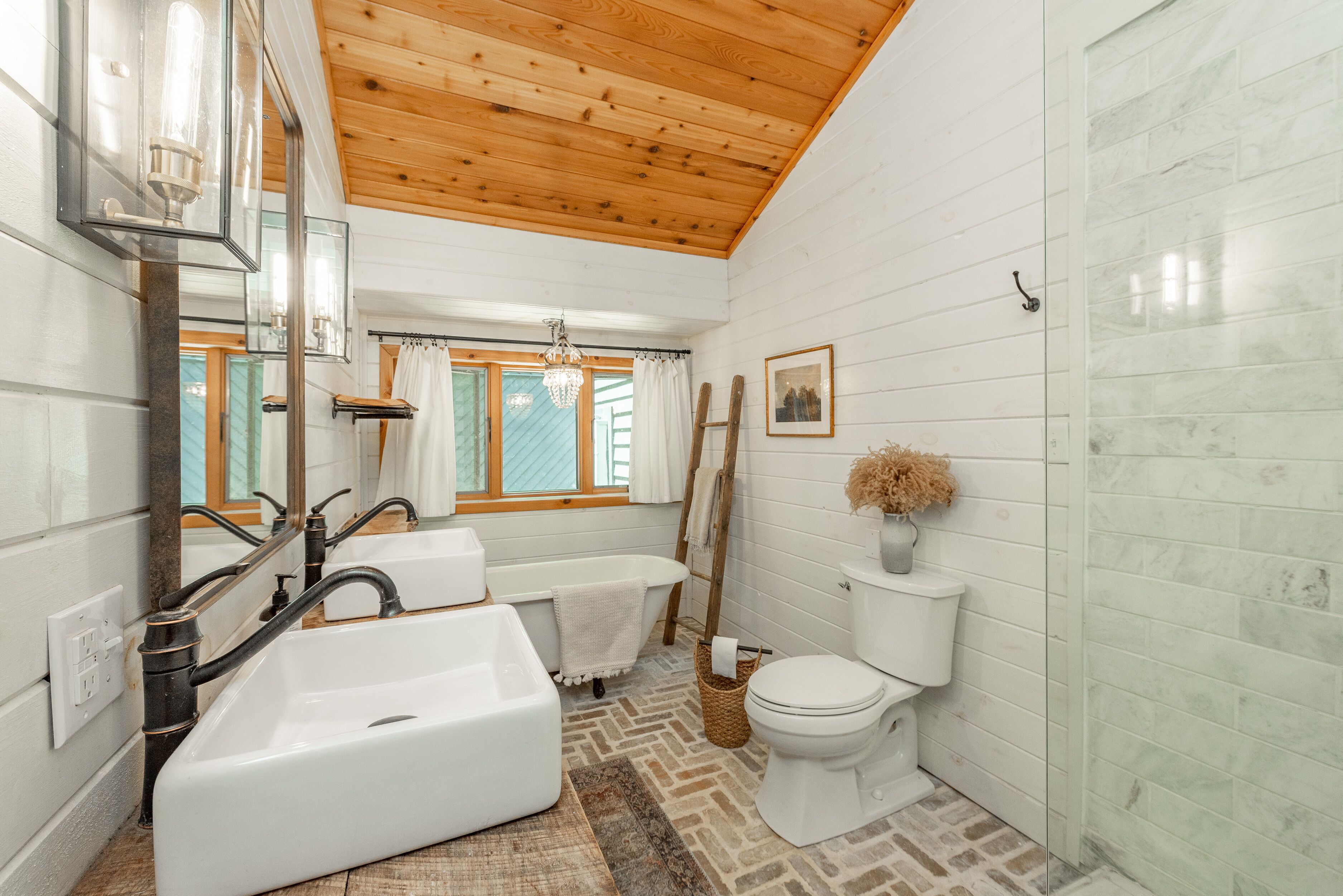 Een badkamer met een houten plafond, witte betegelde wanden, een bad, een toilet en een raam met gordijnen.