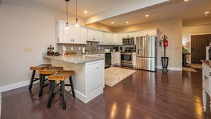 A modern kitchen with white cabinetry, a central island, bar stools, stainless steel appliances, and a wooden floor. - Cozy Deep Creek Cabin w/ Hot Tub & Fireplace (Oakland)