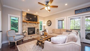 A living room with a stone fireplace, a ceiling fan, a television, and a large window. - Cozy Deep Creek Cabin w/ Hot Tub & Fireplace (Oakland)