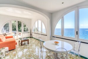A room with a round dining table, clear chairs, a sofa, and large arched windows with a view of the sea.