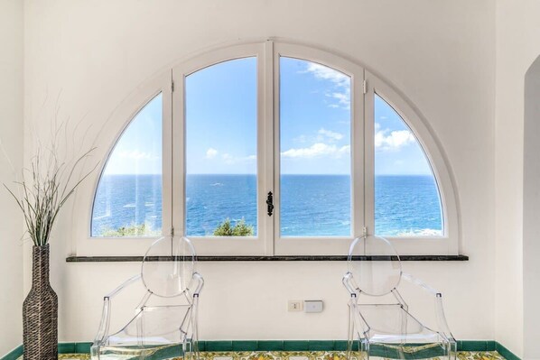 A room with a large arched window overlooking the sea, two clear chairs, and a vase with plants.