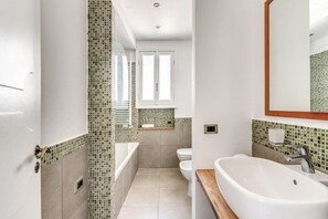 Une salle de bain équipée d’une baignoire, de toilettes et d’un lavabo, avec des murs recouverts de mosaïques et un plan de travail en bois.