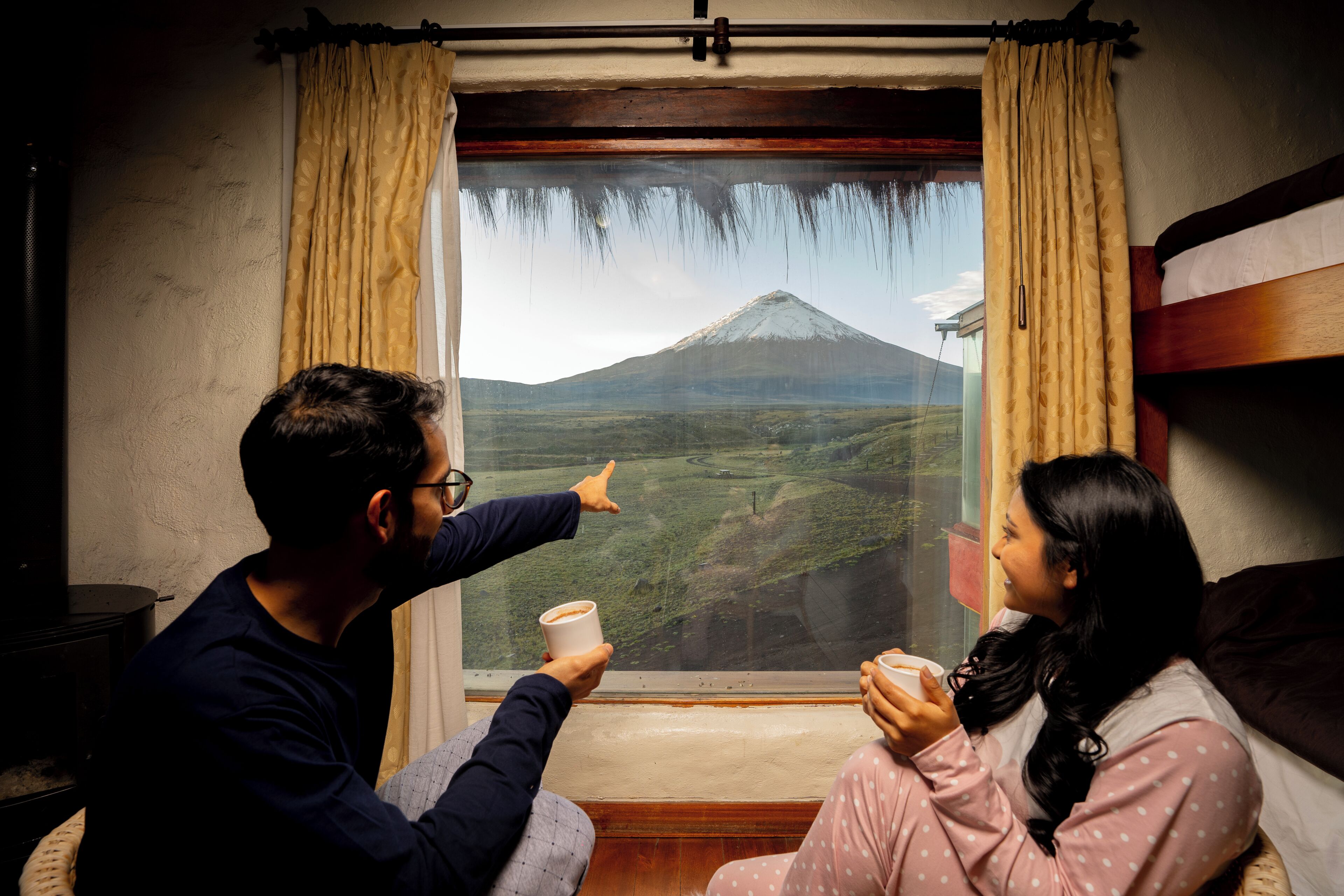 A room with a large window, a bed, and two people sitting by the window.