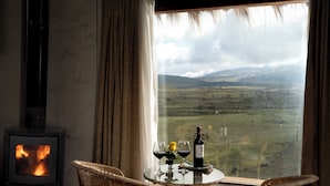 Una habitación con chimenea, una mesa con vasos y una botella, y vista a un paisaje a través de una ventana.