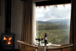 A room with a fireplace, a table with glasses and a bottle, and a view of a landscape through a window.
