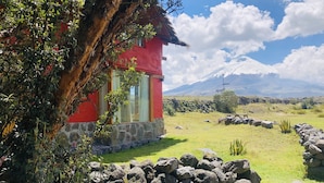 Un edificio rojo con base de piedra, rodeado de un muro de piedra y vegetación, con una montaña al fondo.