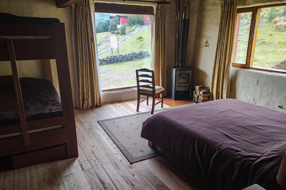 Ein Zimmer mit Etagenbett, einem Sessel, einem Bett mit lila Bettwäsche, einem Kamin und Blick auf ein rotes Haus sowie Grünflächen.