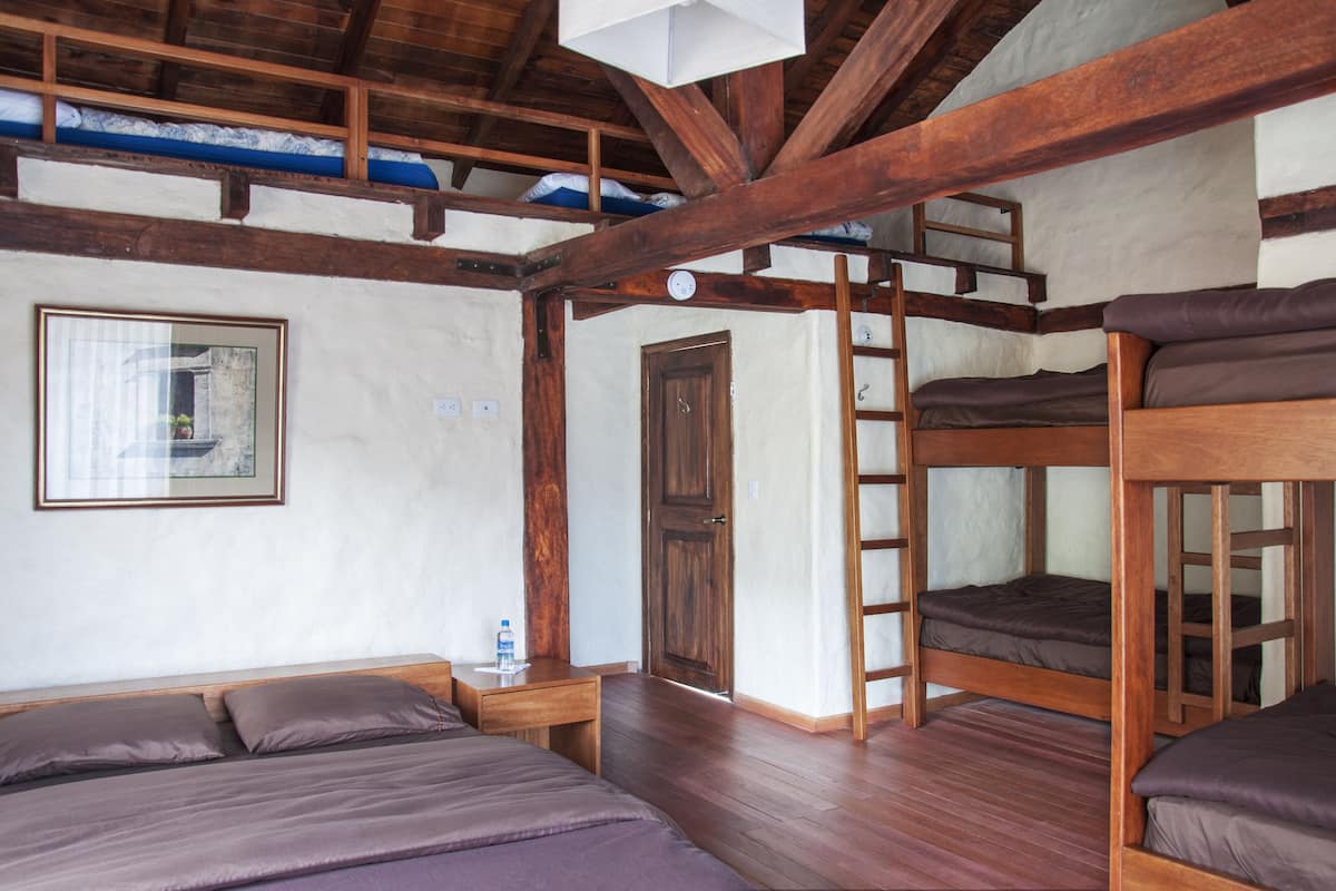 Ein Zimmer mit Etagenbetten, einer Holzleiter, einem kleinen Tisch mit einer Wasserflasche und einem gerahmten Bild an der Wand.