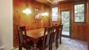 A wooden dining room with a large table and chairs, a painting on the wall, and a door leading outside. - Stairway to Haven ~ Family Cabin in Monte Rio (Monte Rio)