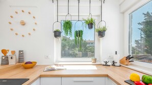 A modern kitchen with a wooden countertop, white cabinets, a window with hanging plants, and a view of the city. - Villa Molaris (Preko)