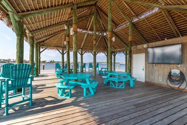 A covered deck with turquoise chairs and picnic tables, a TV screen, and a view of the water.