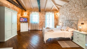 A bedroom with a wooden floor, a stone wall, a bed with white linens, a TV on a white cabinet, and a stone fireplace.